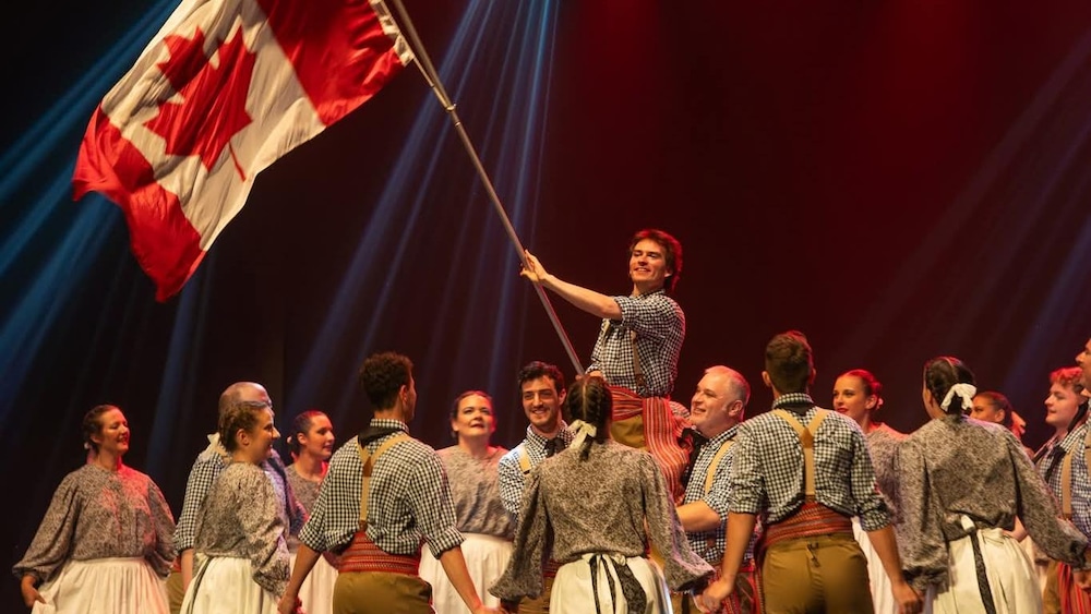 Les danseurs et danseuses de la troupe sont sur scène. Deux personnes en soulèvent une autre qui brandit un immense drapeau du Canada. Les autres membres, main dans la main, forment un cercle autour.