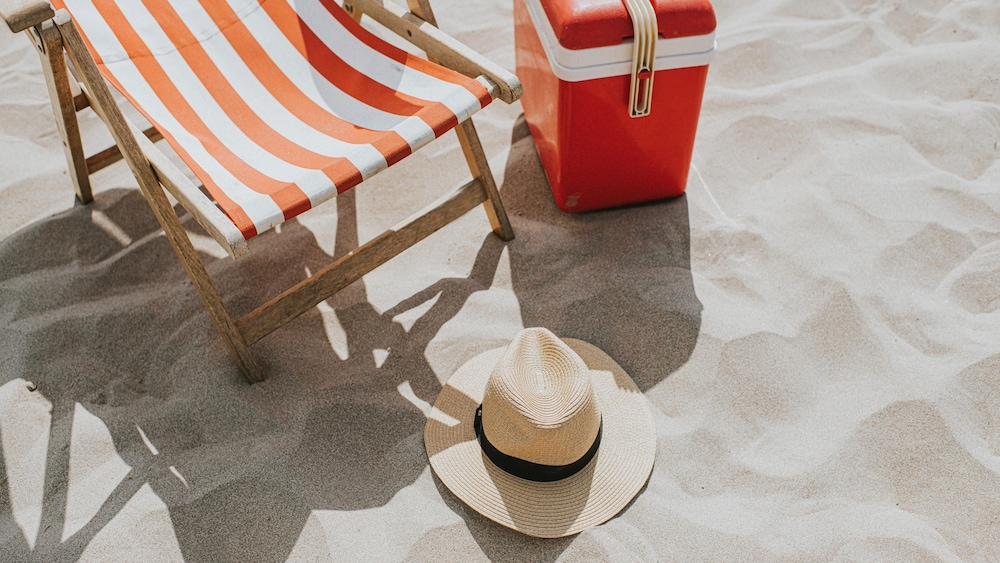 Un chapeau de paille, une glacière et une chaise de plage sur une plage de sable fin. 