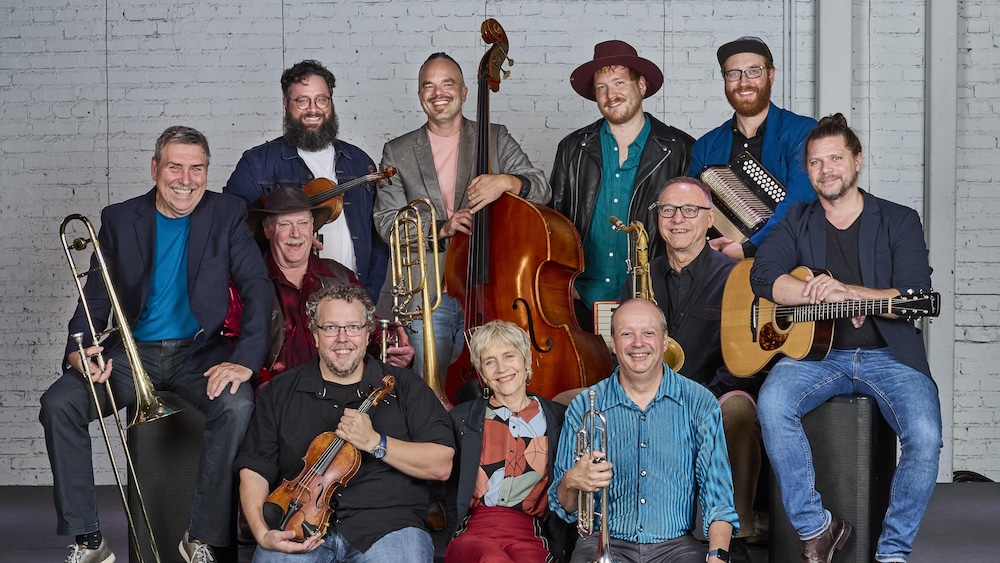 Un groupe de musiciens composé de 10 hommes et une femme sourient en tenant leurs instruments en mains (cuivres, guitare, accordéons, violon, contrebasse).