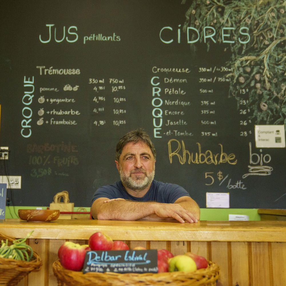 Un homme derrière un comptoir de pommes et de fleurs d'ail. Derrière lui, un menu de jus et cidres.