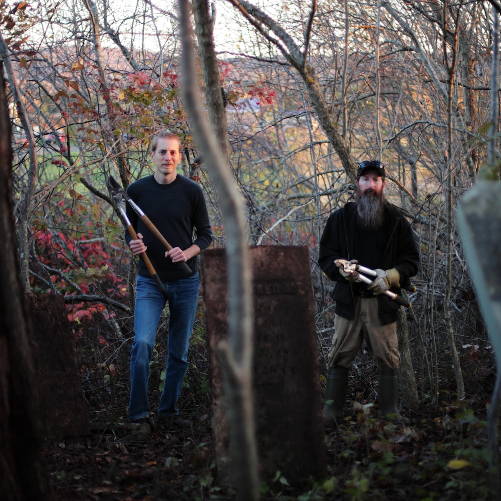 Deux hommes debout dans un cimetière tiennent dans leurs mains des outils de jardin.