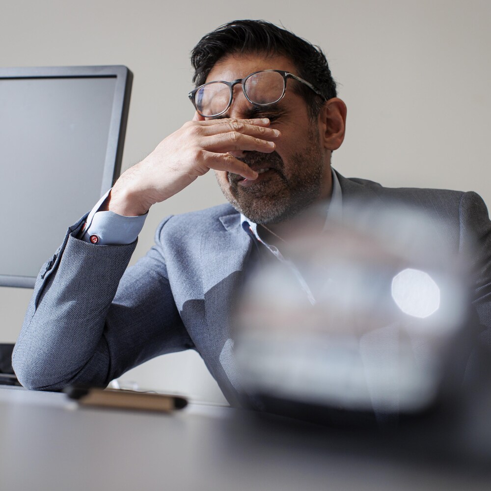 Un homme assis à une table en tenant sa main près de ses yeux en dessous de ses lunettes.