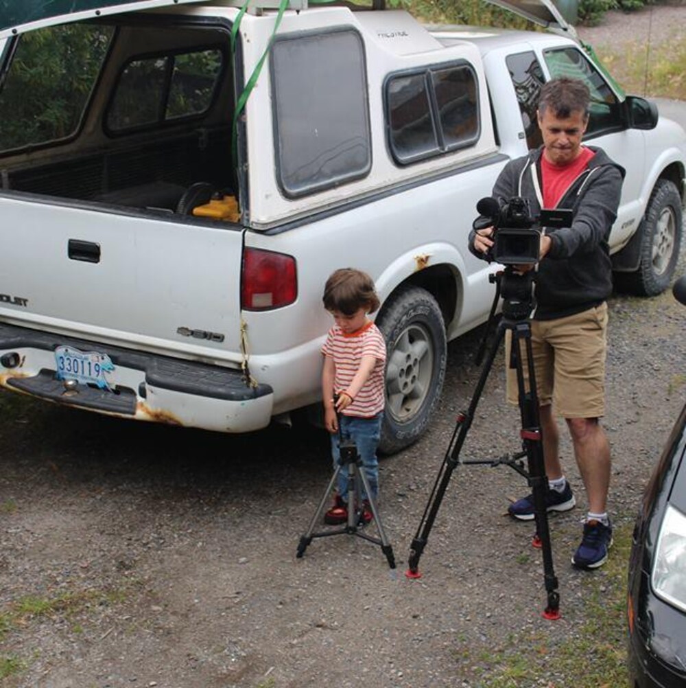 Un enfant manipule un trépied pendant que le réalisateur, à côté de lui, ajuste sa caméra sur un autre trépied. Ils sont dehors, à côté des voitures, dont une camionnette avec un canot sur le toit.