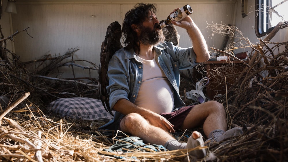 Portrait de Ryan Doucette dans un gros nid d'oiseau, buvant de la bière.