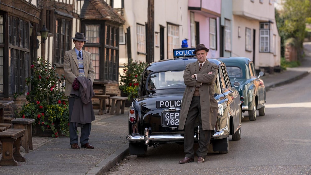 Deux détectives anglais autour de voitures patrouilles anciennes dans la rue d'un village de campagne. 