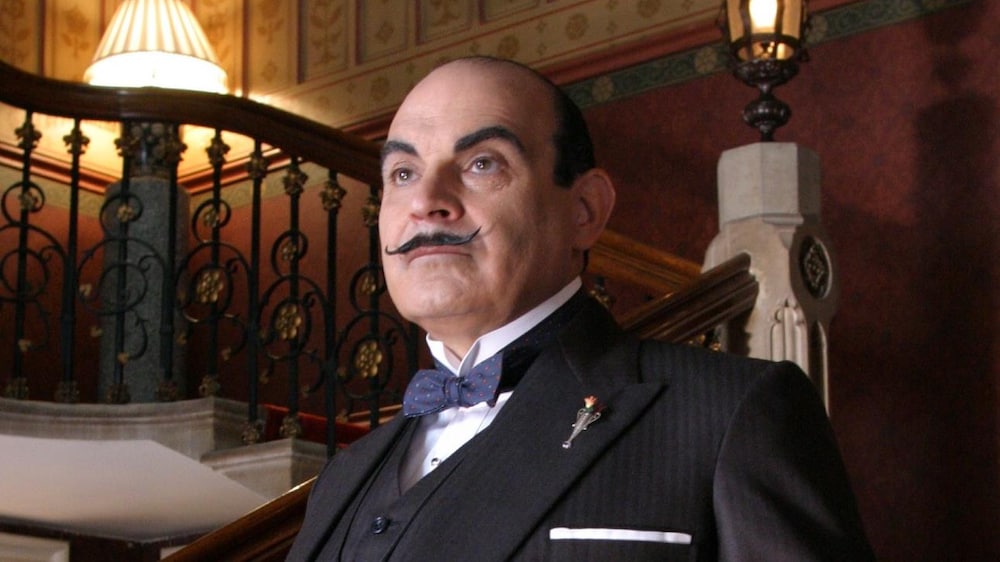 Hercule Poirot (David Suchet) se tient debout devant un somptueux escalier.