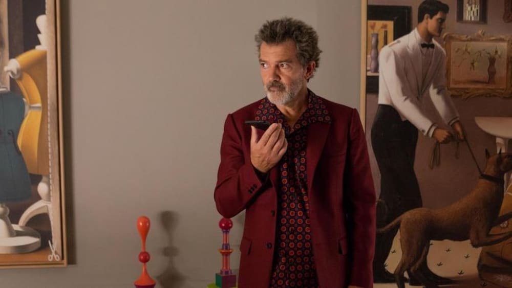 Un homme (Antonio Banderas) tient un téléphone dans ses mains dans une pièce aux murs ornés de tableaux.