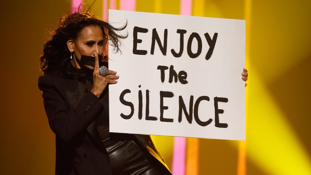 L'animatrice est sur scène, bâillonnée, et elle tient une pancarte sur lequel est écrit «Enjoy the silence».