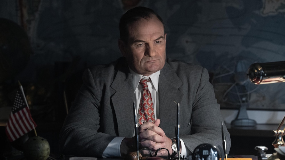 Chris Bauer est assis à son bureau, un mini drapeau des États-Unis devant lui, affichant un regard sévère et perdu dans le vide. 