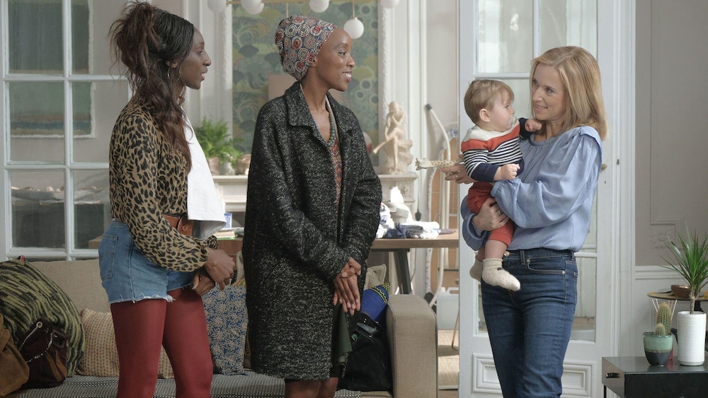 K-Films Amérique Trois femmes, dont l'un tient un bébé dans les bras, dans un salon.