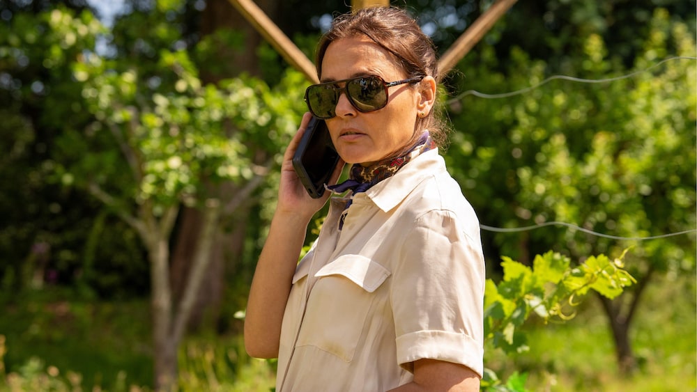 Une femme dans tient un téléphone à l'oreille, dans un décor de campagne.