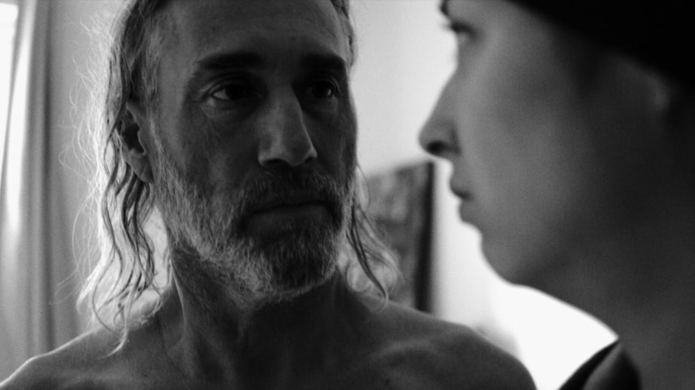 En noir et blanc, un homme torse nu (Roy Dupuis) regarde une jeune femme.