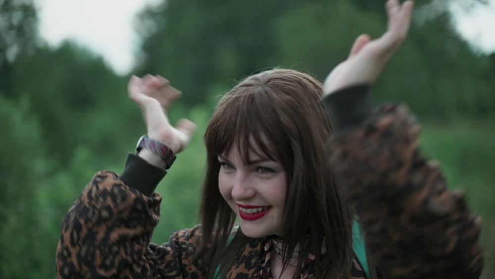 Une jeune fille (Kelly Delpeault) souriante, les bras levés au-dessus d'elle, dans la nature.