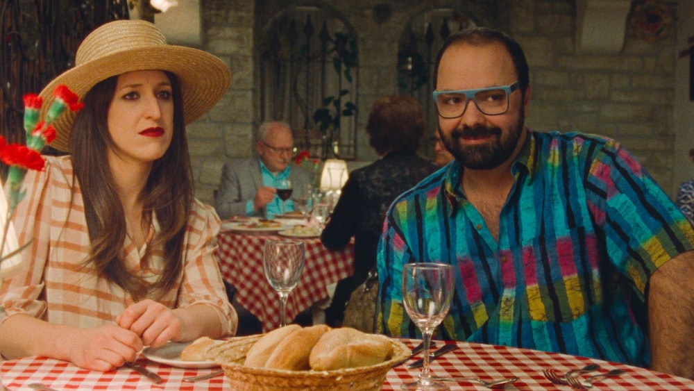 Une femme avec un chapeau (Anne-Élisabeth Bossé) et une homme portant une chemise colorée (Mani Soleymanlou) attablés à un restaurant.