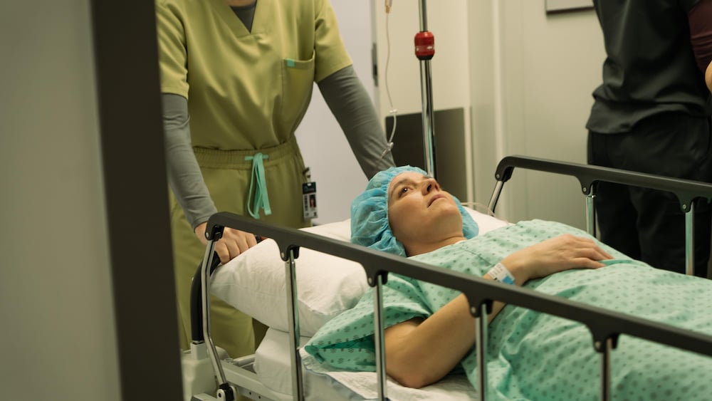Marie-Ève Perron est sur une civière avec une blouse d'hôpital et un bonnet sur les cheveux.