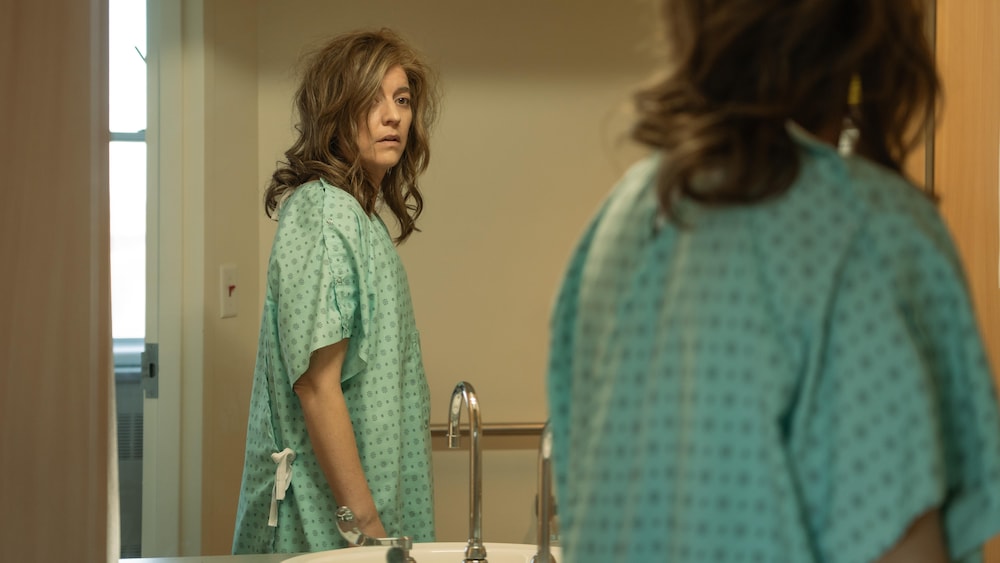Marie-Ève Perron est debout dans une blouse d'hôpital et regarde sa poitrine plate dans le miroir.