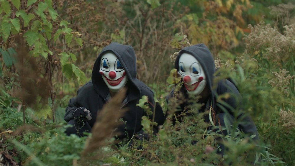 Radio-Canada Catherine et Tonya, toutes deux vêtues de vestes noires et portant des masques de clown, se cachent dans une forêt.