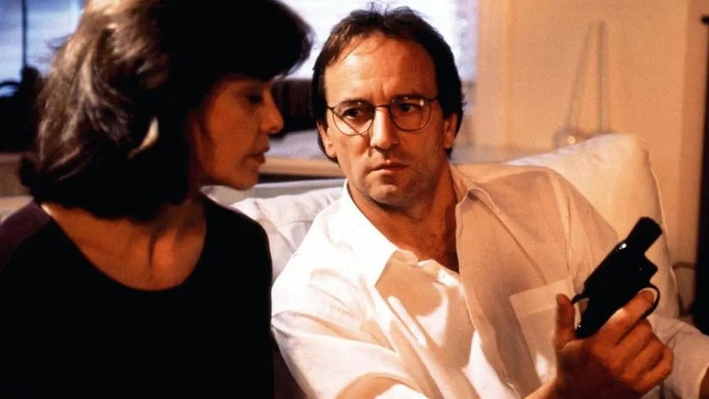 Astral Films Une femme assise sur un canapé à côté d'un homme (Michel Côté) qui la regarde et qui tient un pistolet.