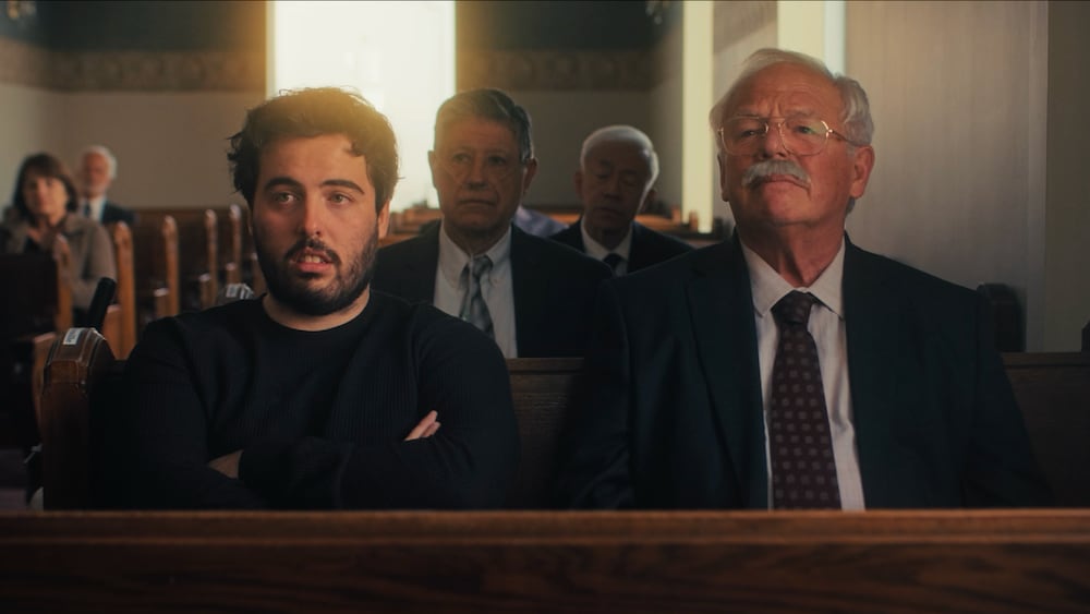 Les deux personnages sont assis sur un banc dans un salon funéraire.