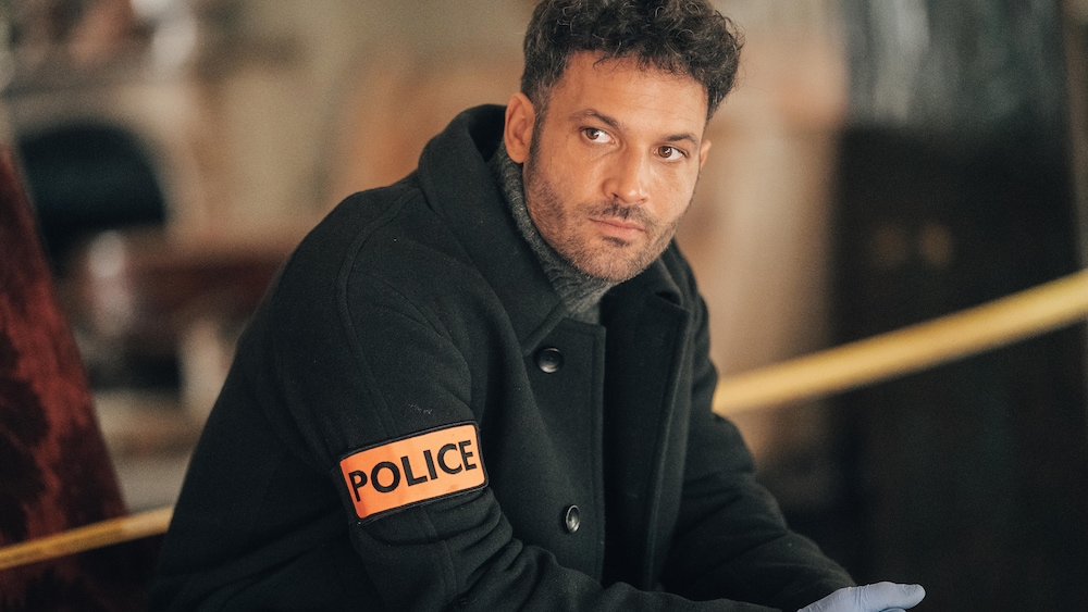 © caroline dubois - ftv - jla Nicolas est sur une scène de crime. Il porte un mateau qui affiche une étiquette avec le mot police et des gants chirurgicaux.