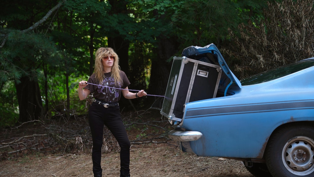 karolina wojtasik/peacock / Peacock La femme est debout derrière une voiture bleue. Le coffre est ouvert et un coffre noir en sort.