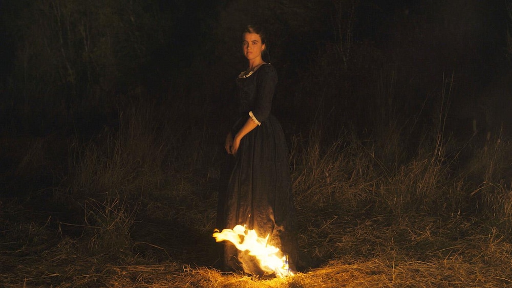 Une jeune femme en robe d'époque devant un feu dans un champ, la nuit.