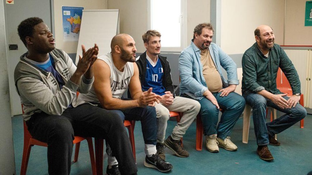 EyeSteel Films Plusieurs hommes assis sur des chaises en plastique dans un local rient.