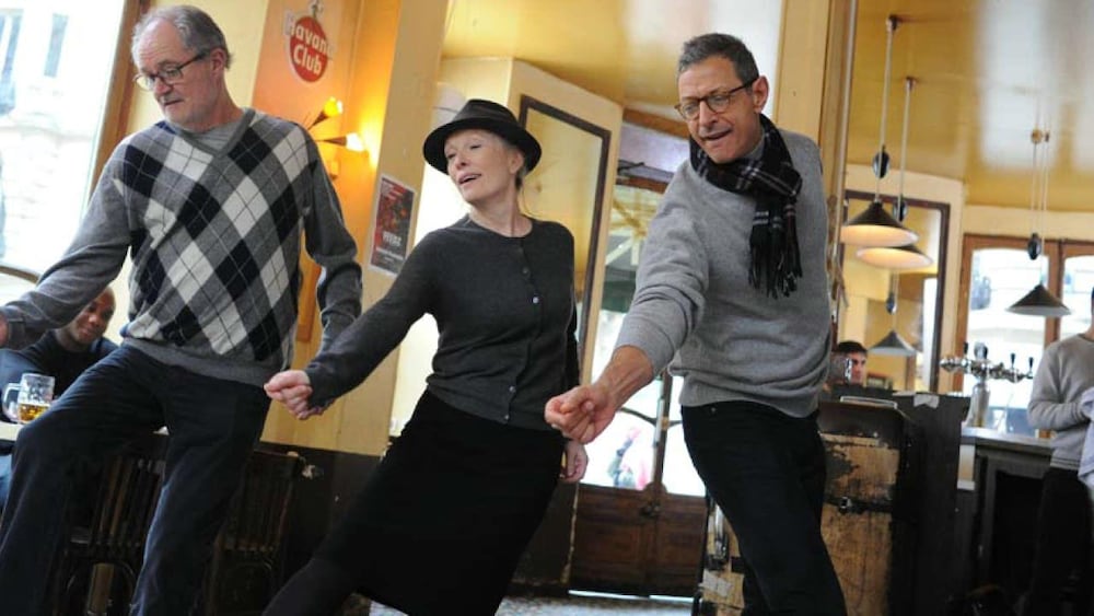 Dans un café parisien, deux hommes et une femme portant un chapeau esquissent un pas de danse.