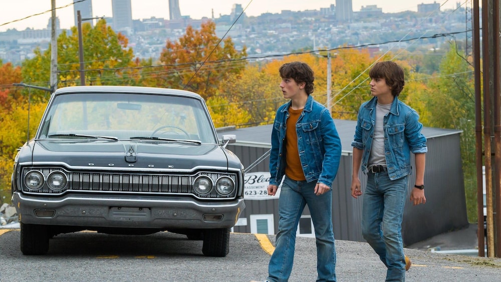 Deux jeunes hommes habillés de jeans traversent la rue, devant une vieille voiture.