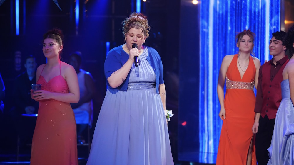 Plan moyen de Coco Belliveau, en robe de bal bleu pâle et une veste de cardigan assortie, chantant avec un micro dans une ambiance intimiste sur une scène éclairée de lumières bleues.