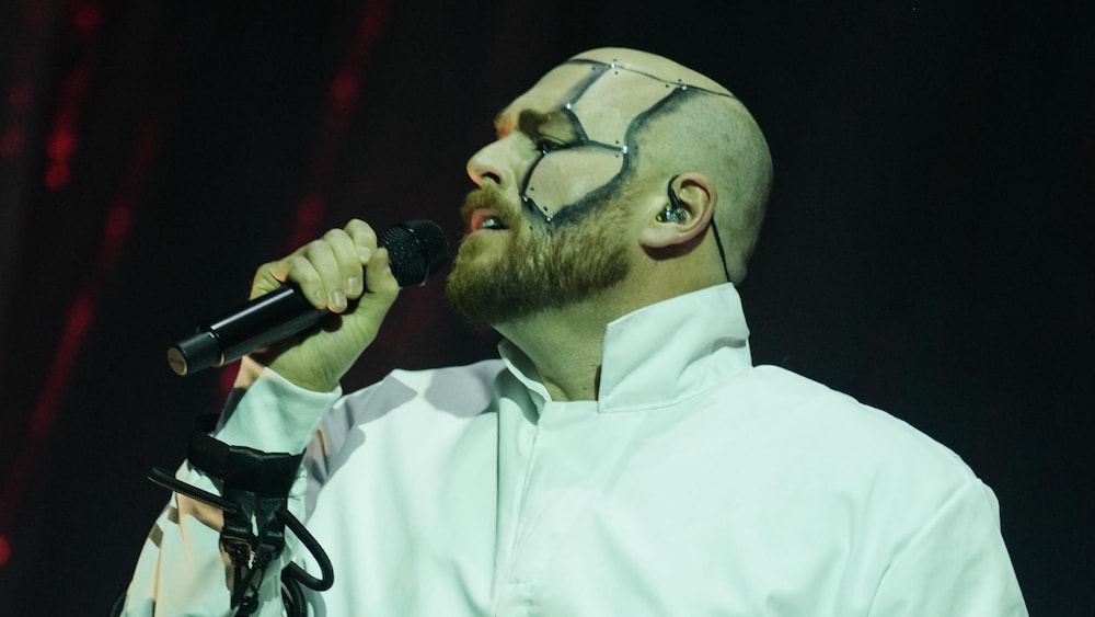 Constant Bernard, en costume blanc avec peinture faciale, chante avec passion, les yeux fermés.