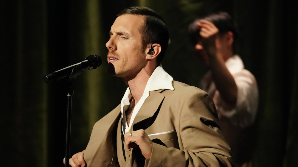 L'animateur Pascal Morrissette chante avec intensité, un micro à la main, sur une scène au rideau vert drapé, portant un costume marron clair et une chemise blanche ouverte.