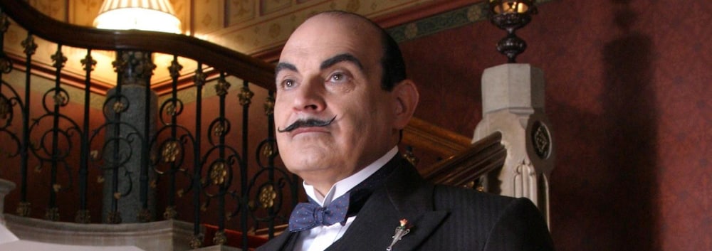 Hercule Poirot (David Suchet) se tient debout devant un somptueux escalier.