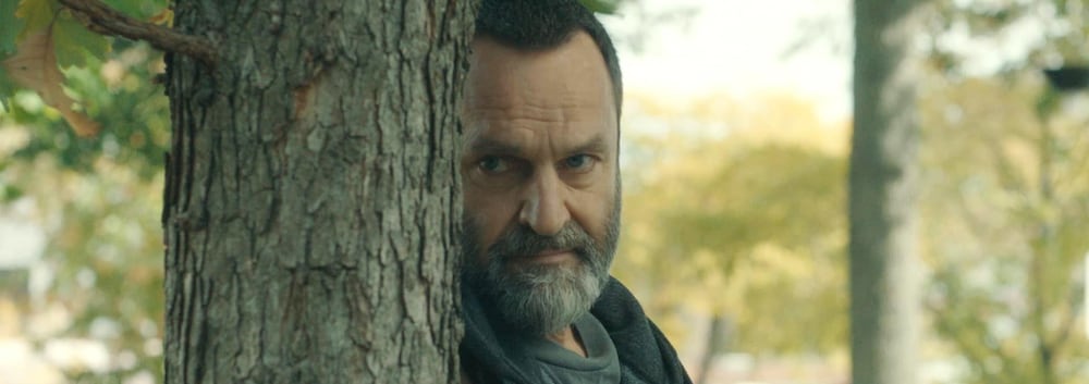 Larry (Benoît Gouin) fait de la surveillance planqué derrière un arbre.