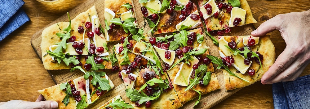 Des mains prennent une portion de pain plat coupé, recouvert de fromage brie, roquette et canneberges.