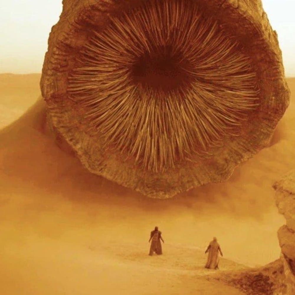 Dans un désert, la bouche d'un immense monstre des sables devant deux silhouettes.