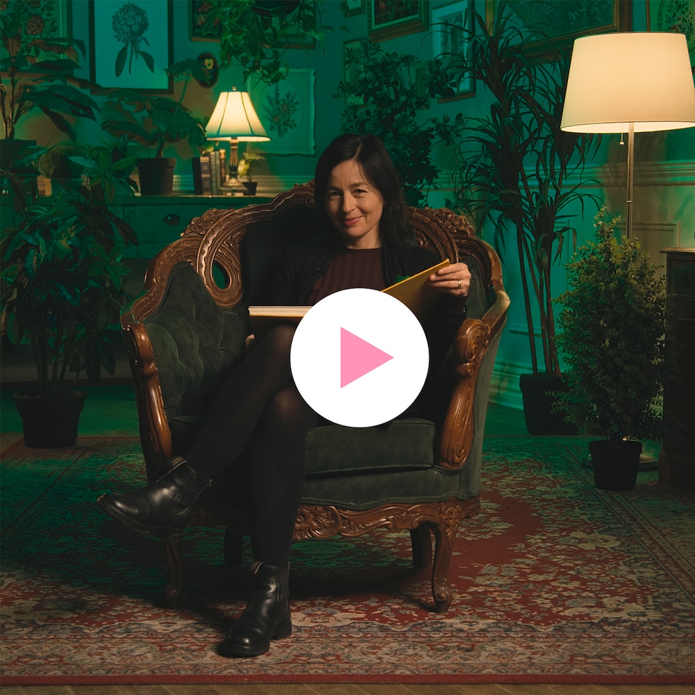 Laure Waridel est assise dans un fauteuil sur un tapis dans une pièce entourée de plantes et de tableaux botaniques.