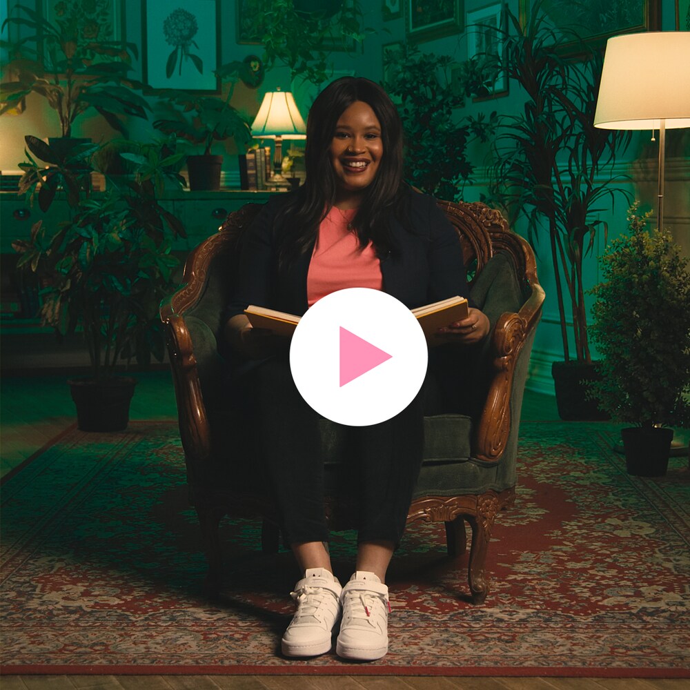Vanessa Destiné est assise dans un fauteuil sur un tapis dans une pièce entourée de plantes et de tableaux botaniques.