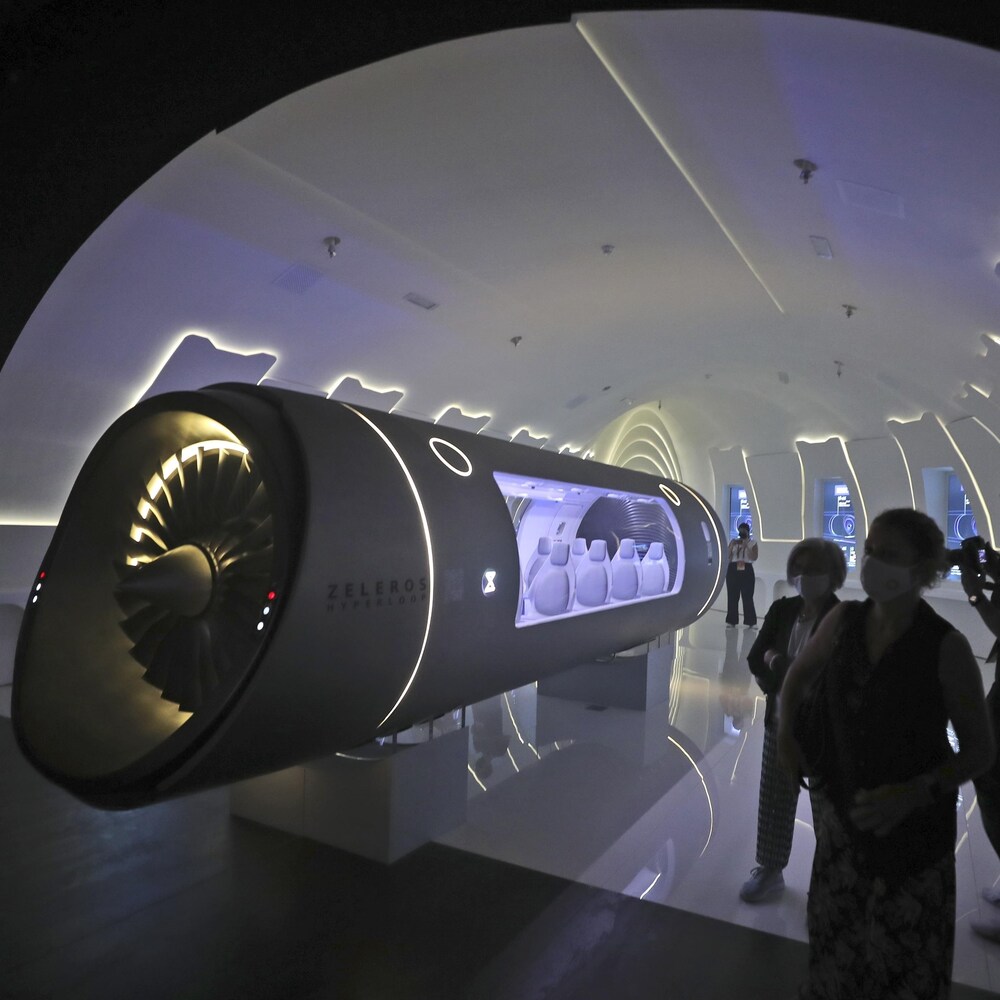 Trois personnes en silhouette observent un véhicule hyperloop dans une salle d'exposition.