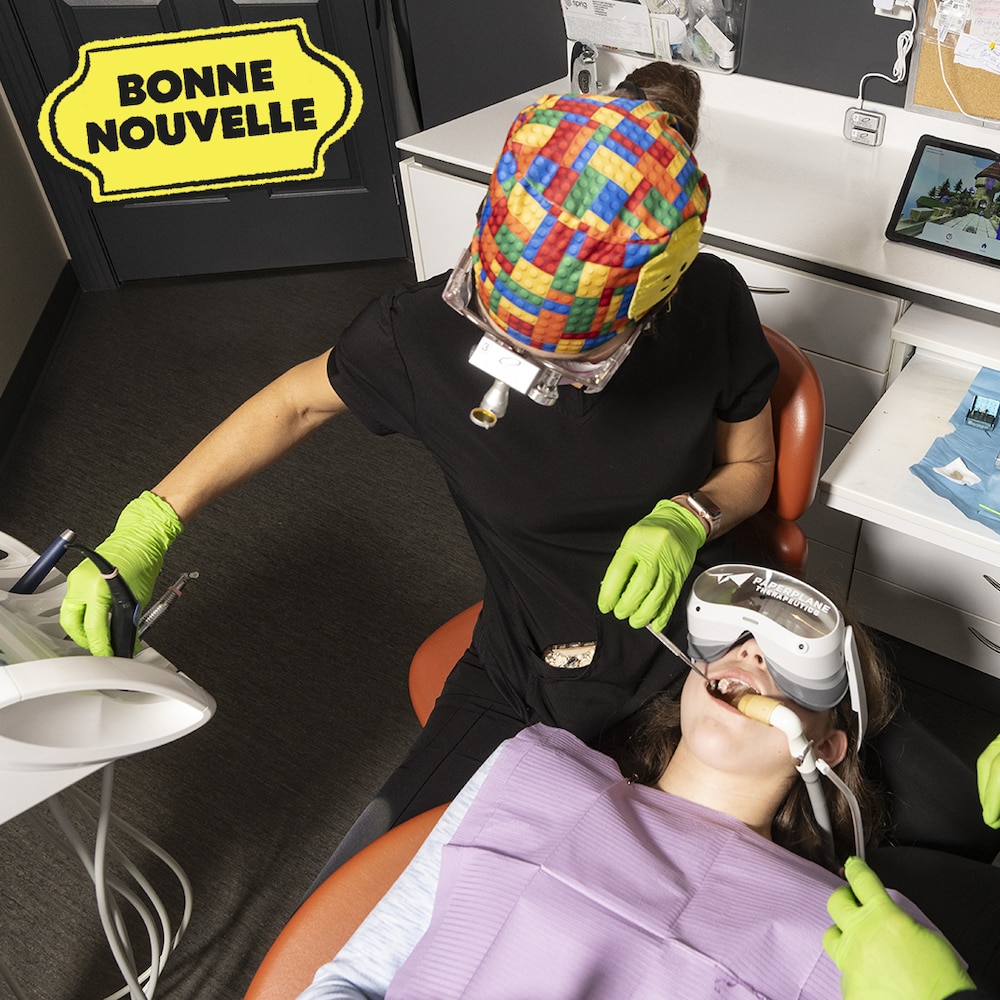 Une jeune fille est assise dans une chaise et porte un casque de réalité virtuelle. Deux professionnelles de la santé dentaire inspectent sa bouche.