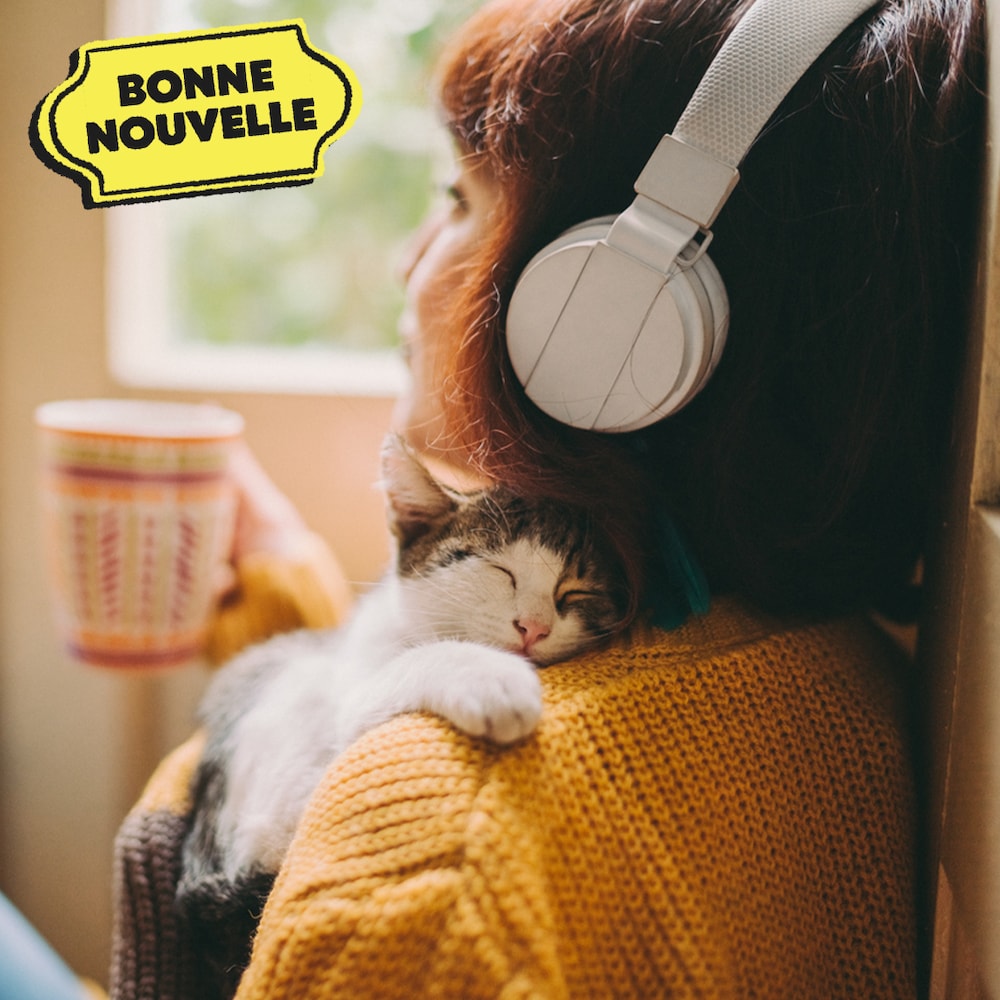 Une jeune femme qui porte des écouteurs est assise avec une tasse et un chat qui dort sur son épaule.