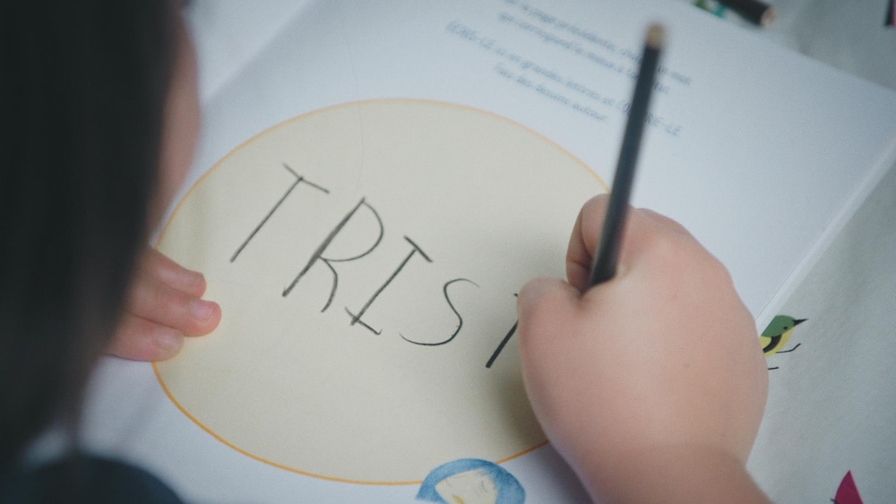 Radio-Canada Un enfant écrit le mot « triste » dans un cahier à colorier.