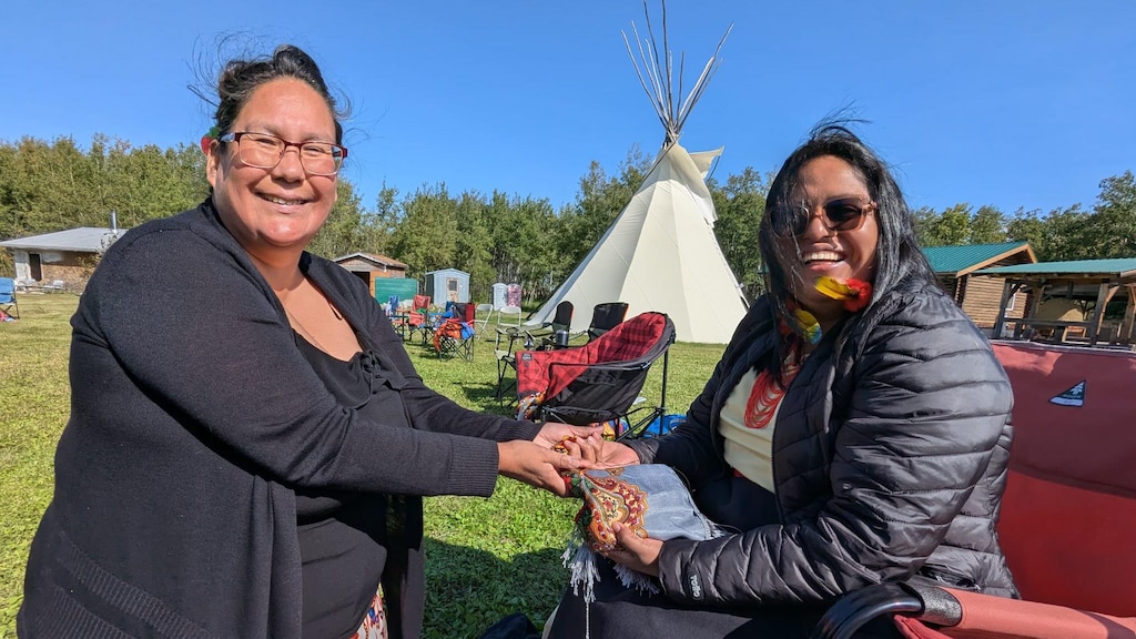 ES: Dos mujeres sonrientes interactúan al aire libre en un día soleado; un tipi blanco al fondo.
FR: Deux femmes souriantes interagissent en plein air par une journée ensoleillée ; un tipi au fond.
EN: Two smiling women interact outdoors on a sunny day, with a white teepee visible in the background.