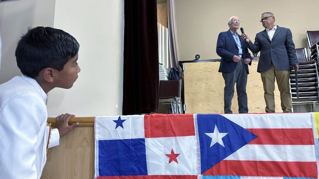 ES: Un niño observa desde abajo a dos hombres hablando por micrófono en el escenario.
FR: Un garçon regarde d'en bas deux hommes parlant au micro sur la scène.
EN: A young boy looks up at two men speaking into a microphone on the stage.