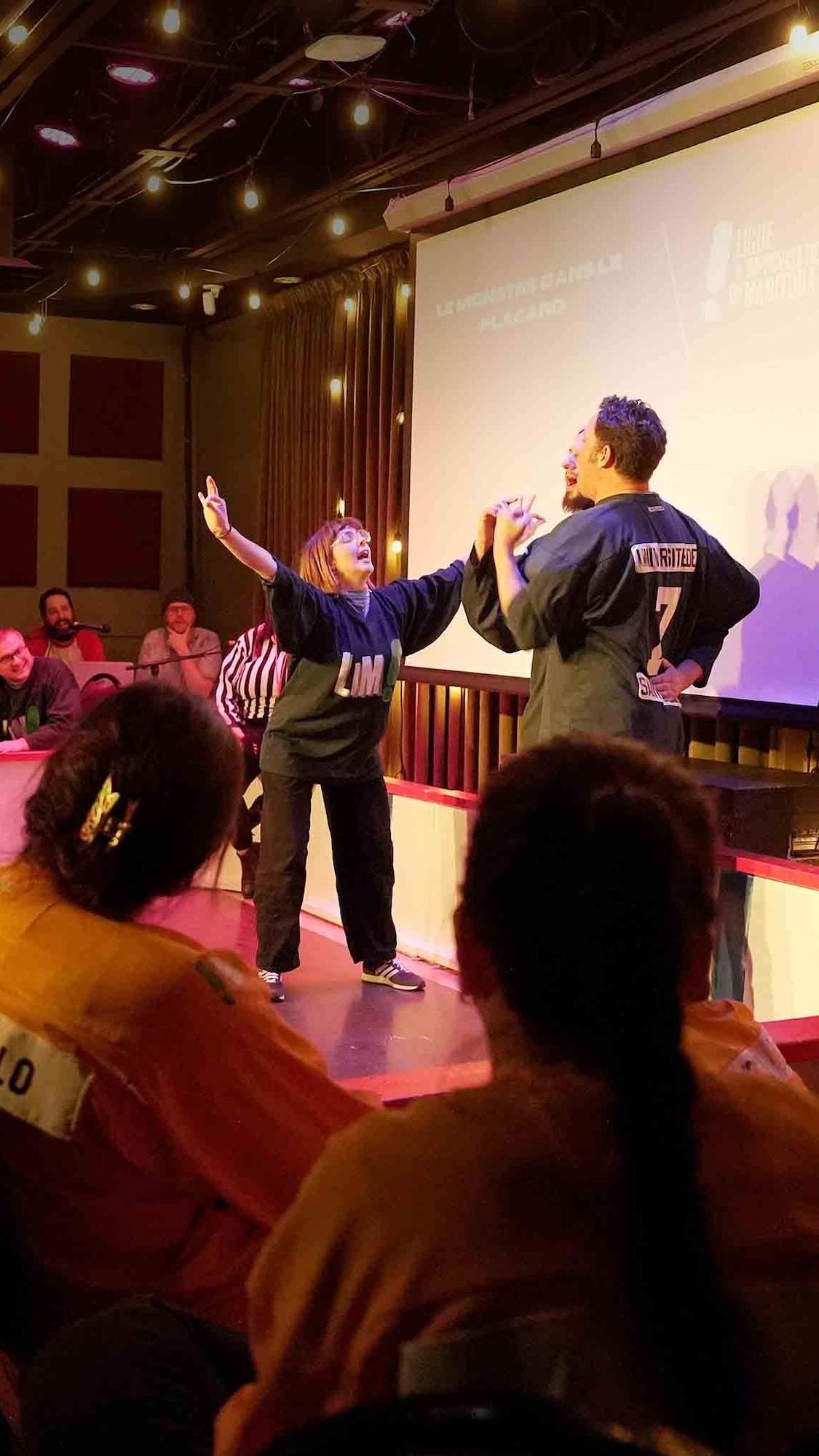 Deux improvisateurs sur une scène sombre, les bras levés en signe de victoire ou de joie intense.