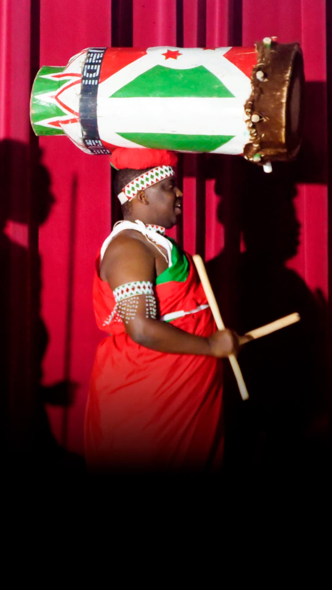 Sur une scène devant un rideau rouge, un homme et un jeune garçon portent des toges rouges et vertes avec des bandeaux à motifs géométriques. L'homme, portant un grand tambour décoré en équilibre sur sa tête, tient des baguettes en bois, tout comme le garçon debout à ses côtés.