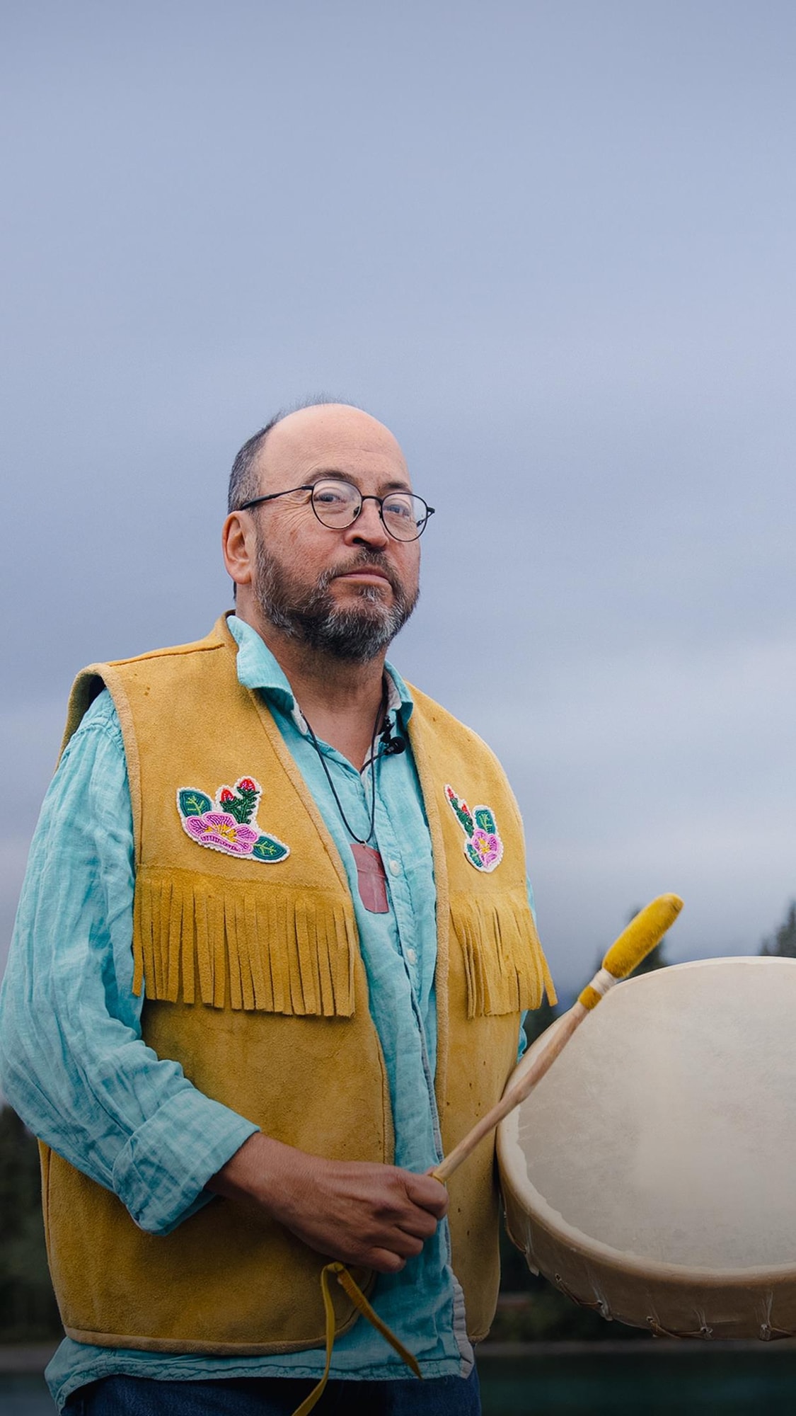 John Fingland, arborant un vêtement traditionnel autochtone et tambour à la main, s’apprête à raconter ses histoires, au bord du lac Kathleen, au Yukon, en août 2026.