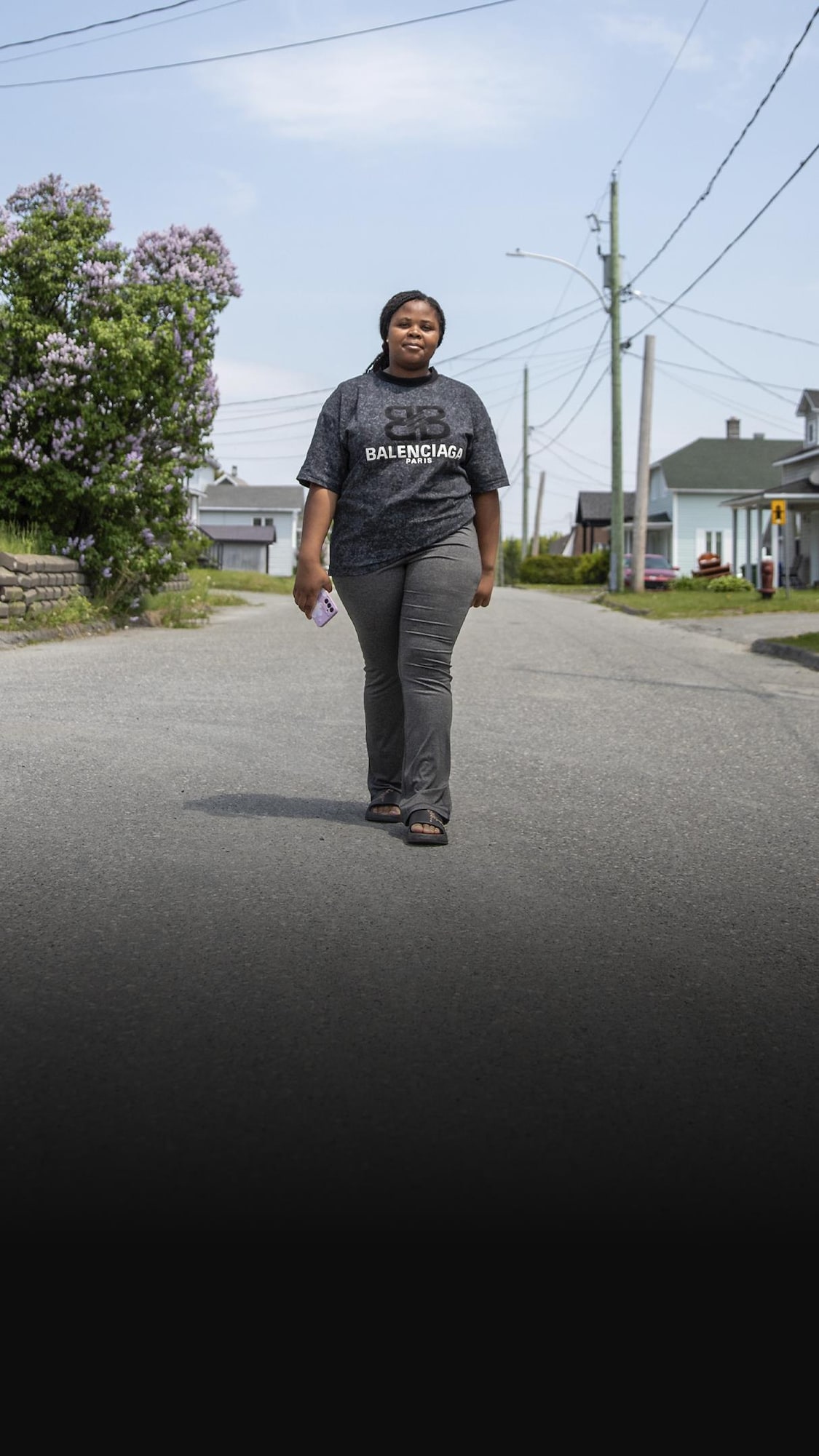 Une jeune femme vêtue d'un t-shirt gris avance au milieu d'une rue résidentielle bordée de maisons.