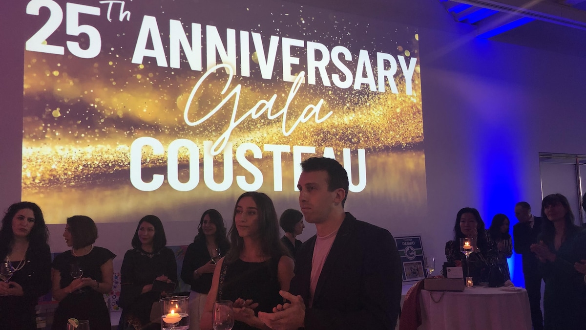 Plusieurs personnes en habits de soirée applaudissent, avec, en arrière-plan, un écran sur lequel il est écrit en anglais Gala du 25e anniversaire de Cousteau.