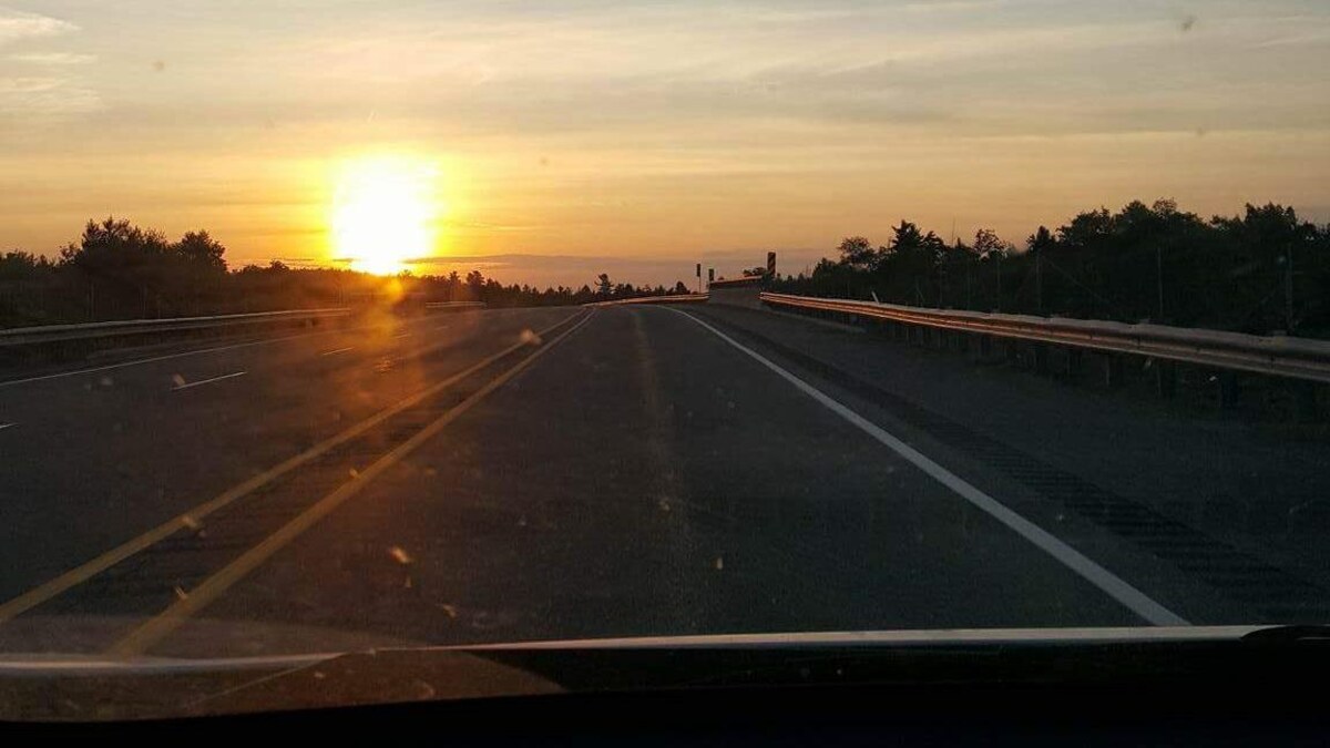 Par le pare-brise, nous voyons un soleil se lever dans un ciel parsemé de quelques nuages.